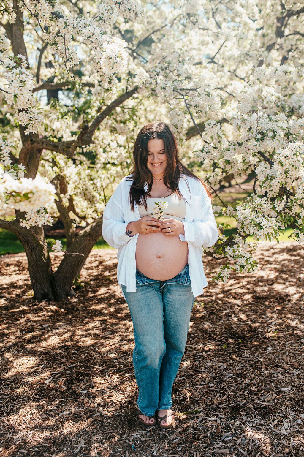 Pregnant person standing under blooming trees, holding belly and smiling outdoors