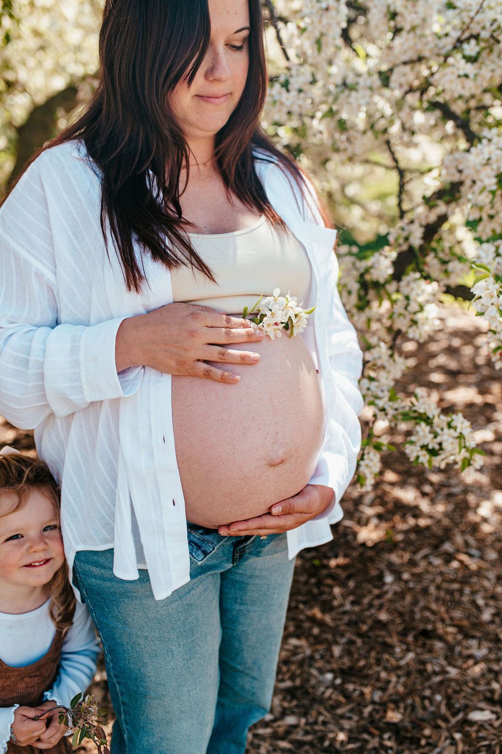 Pregnant woman in white cardigan standing outdoors with a small child beside her