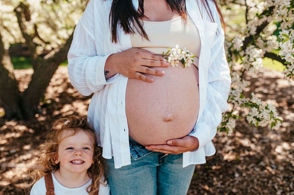 Pregnant person holding belly with a smiling child beside them outdoors among blooming trees