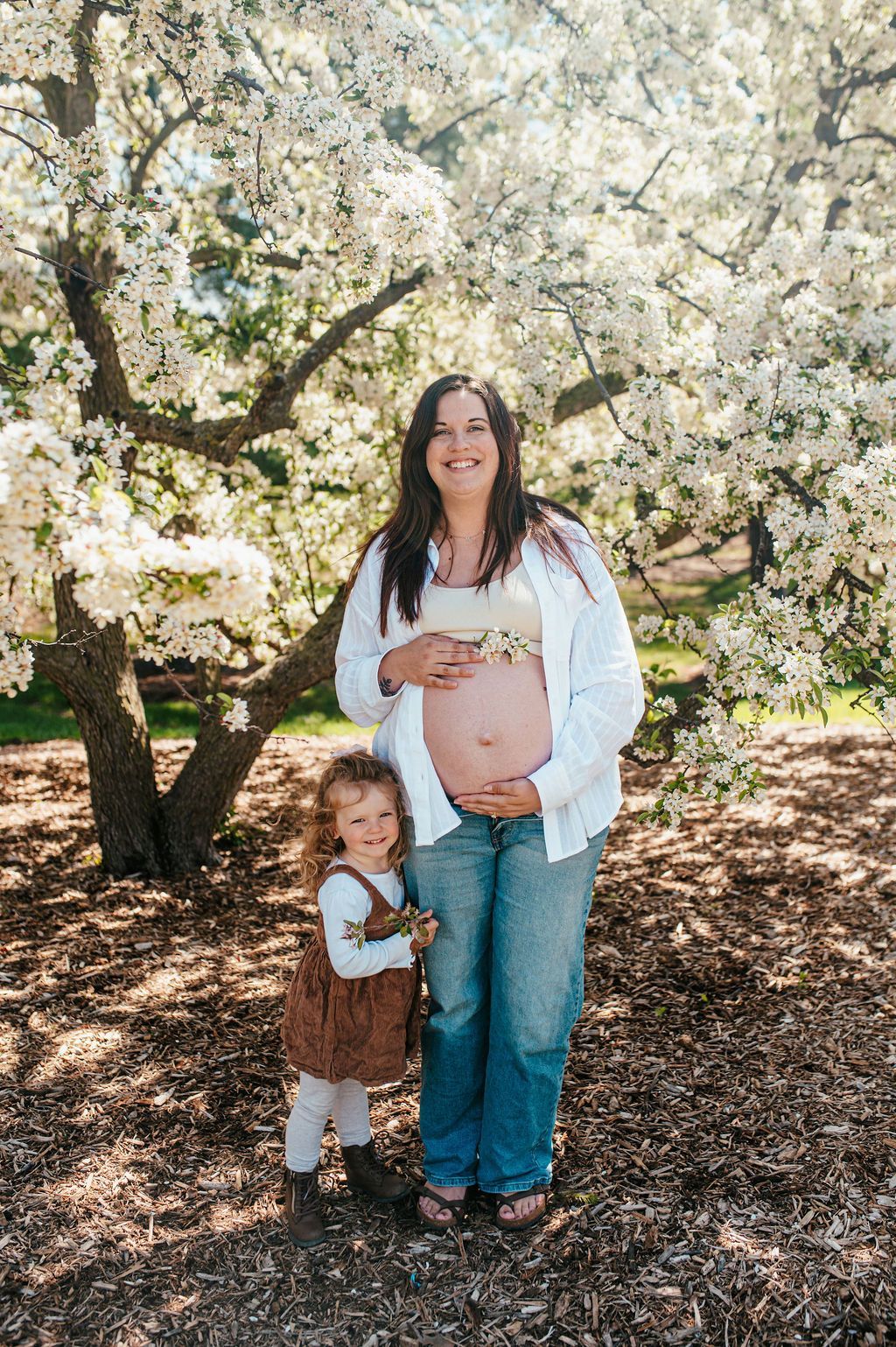 Pregnant person standing with a small child under blooming white blossoms outdoors