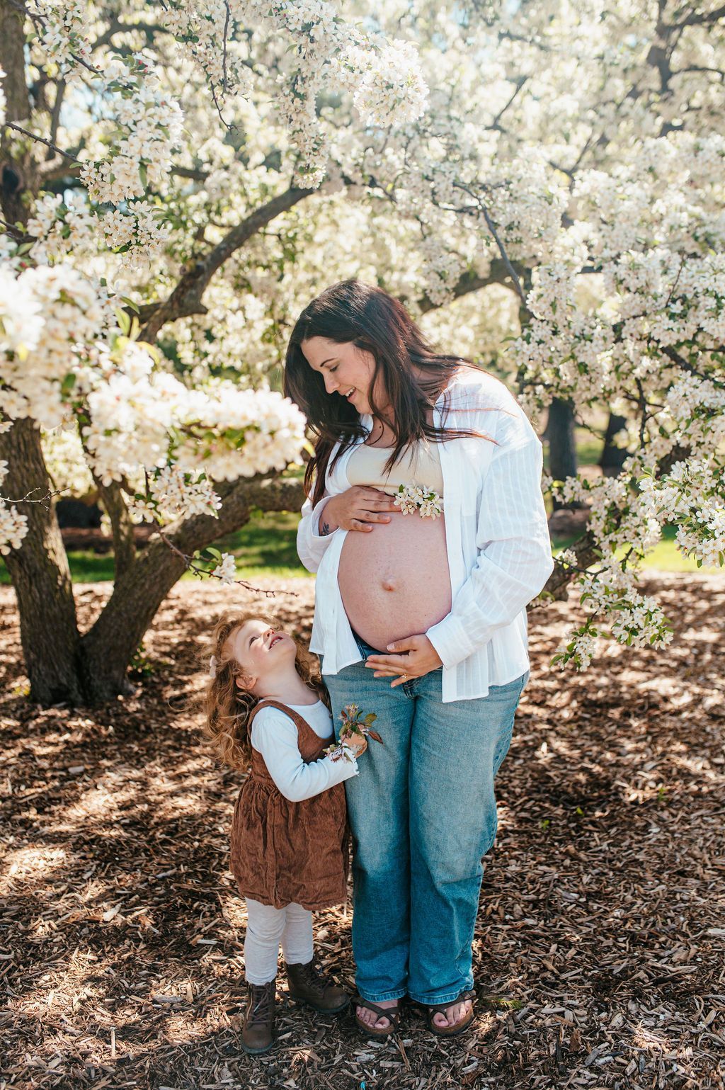 Pregnant woman and child standing under blooming white blossoms in a sunny garden