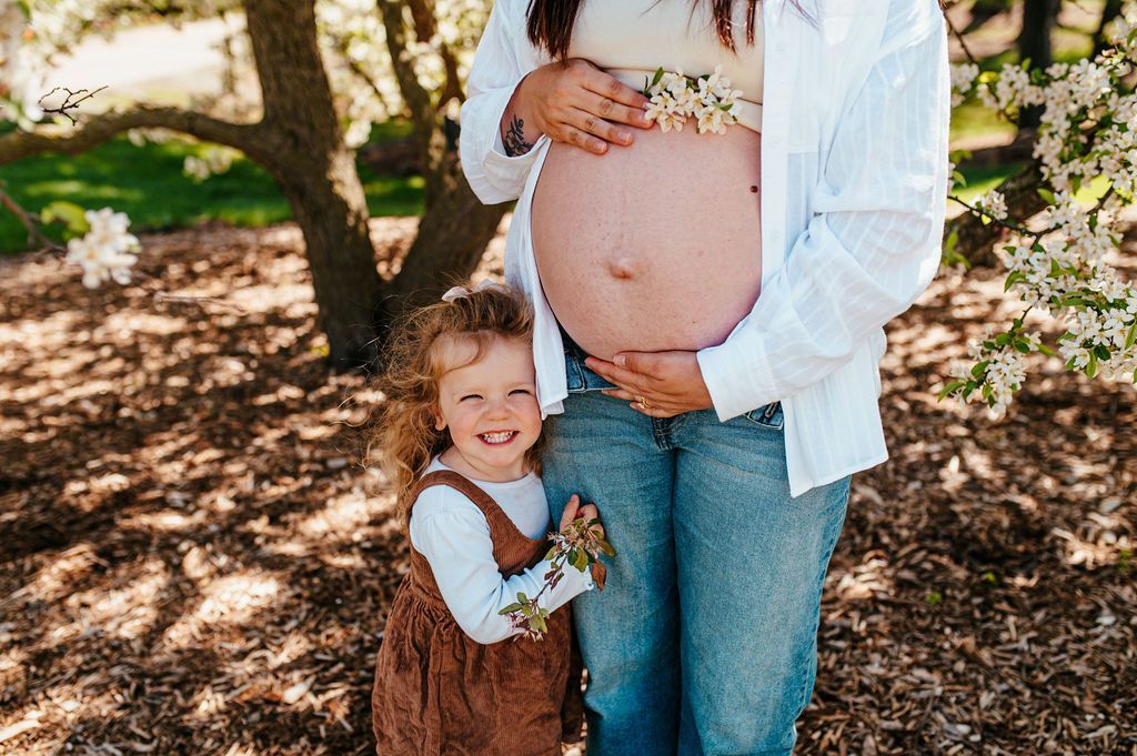 Pregnant person with child in a sunlit wooded area, both wearing casual clothes and smiling