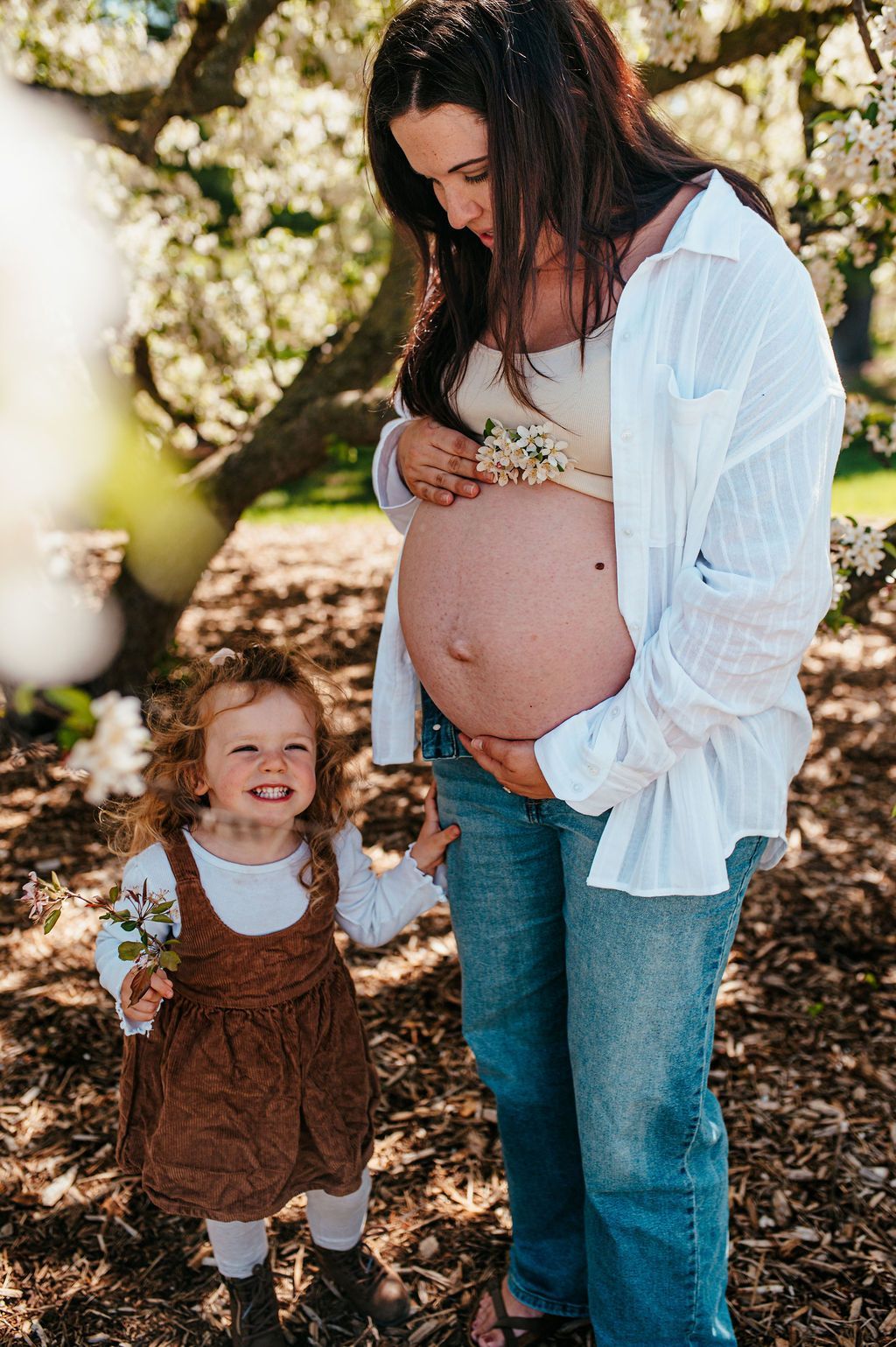 Pregnant woman standing outdoors with a smiling child in brown overalls and white shirt