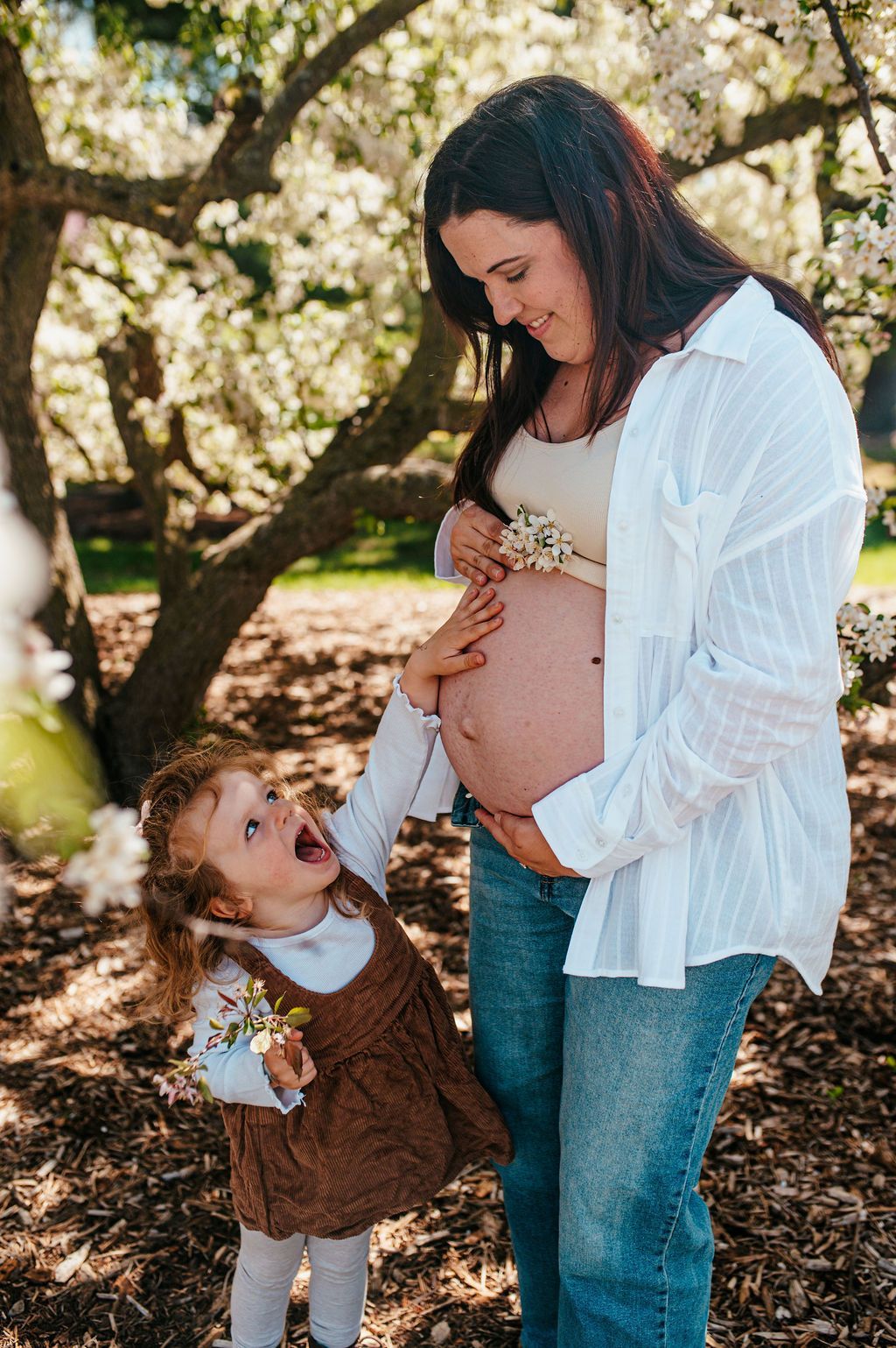 Pregnant woman in a white shirt smiling at a little girl in a brown dress outdoors under blooming trees