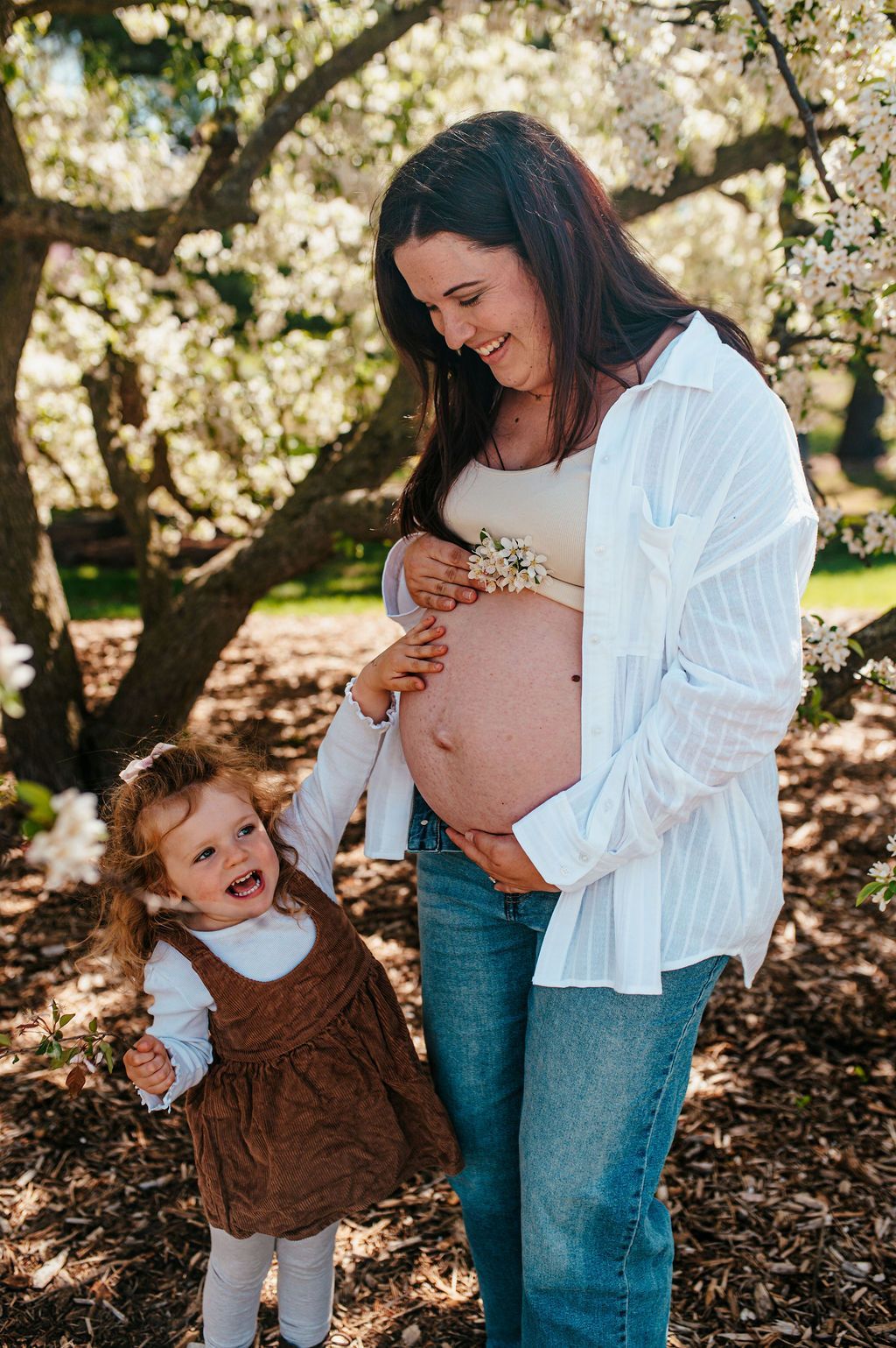 Pregnant woman in white shirt standing with a young child under blooming trees outdoors
