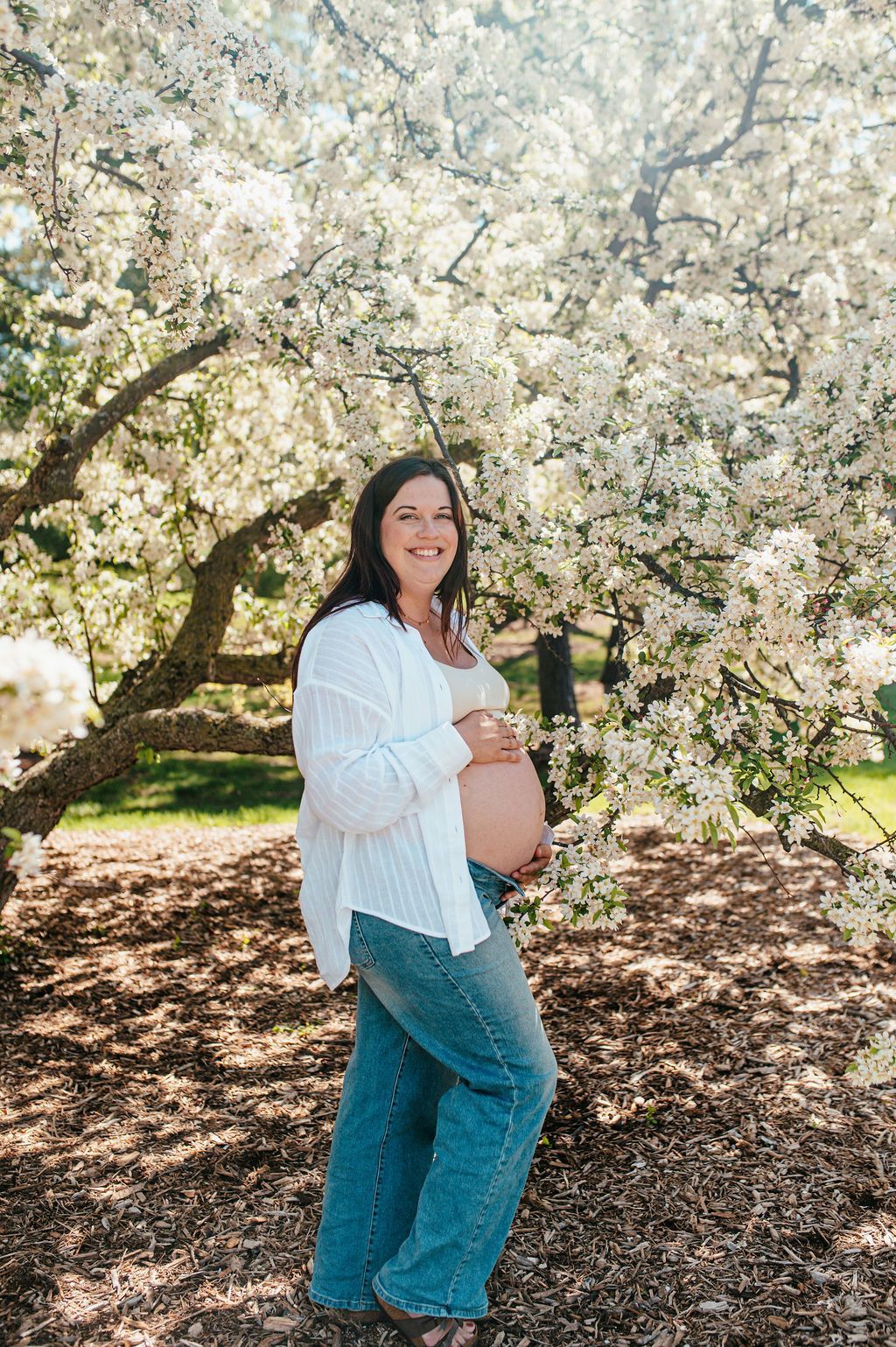 Pregnant woman in a white blouse and jeans standing under blooming white blossoms outdoors