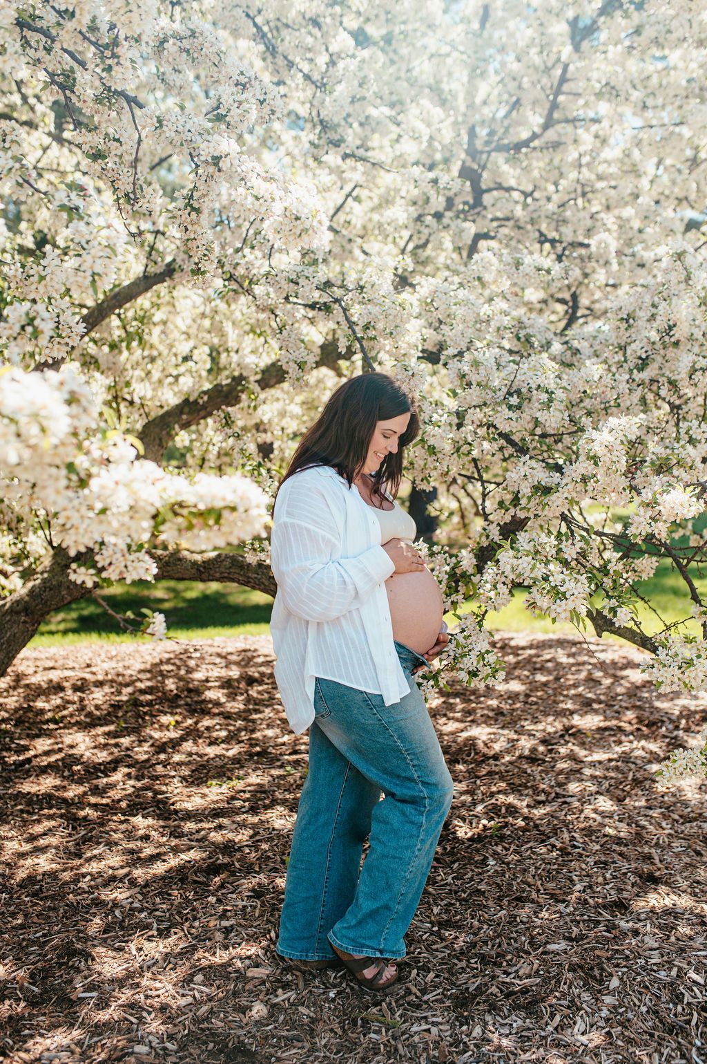 Pregnant person standing under blooming white trees in a sunlit park, hand on belly