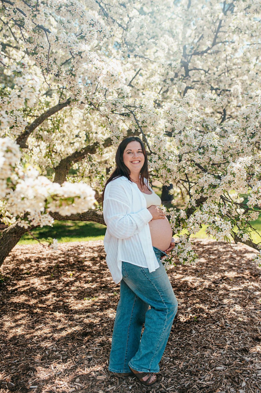 Person standing in front of blooming white trees, smiling in a white top and blue jeans.