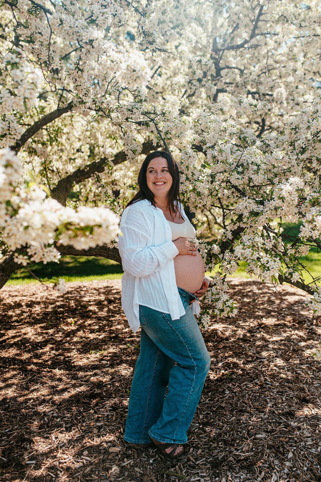 Pregnant person in a white shirt and blue jeans standing beneath blooming trees outdoors