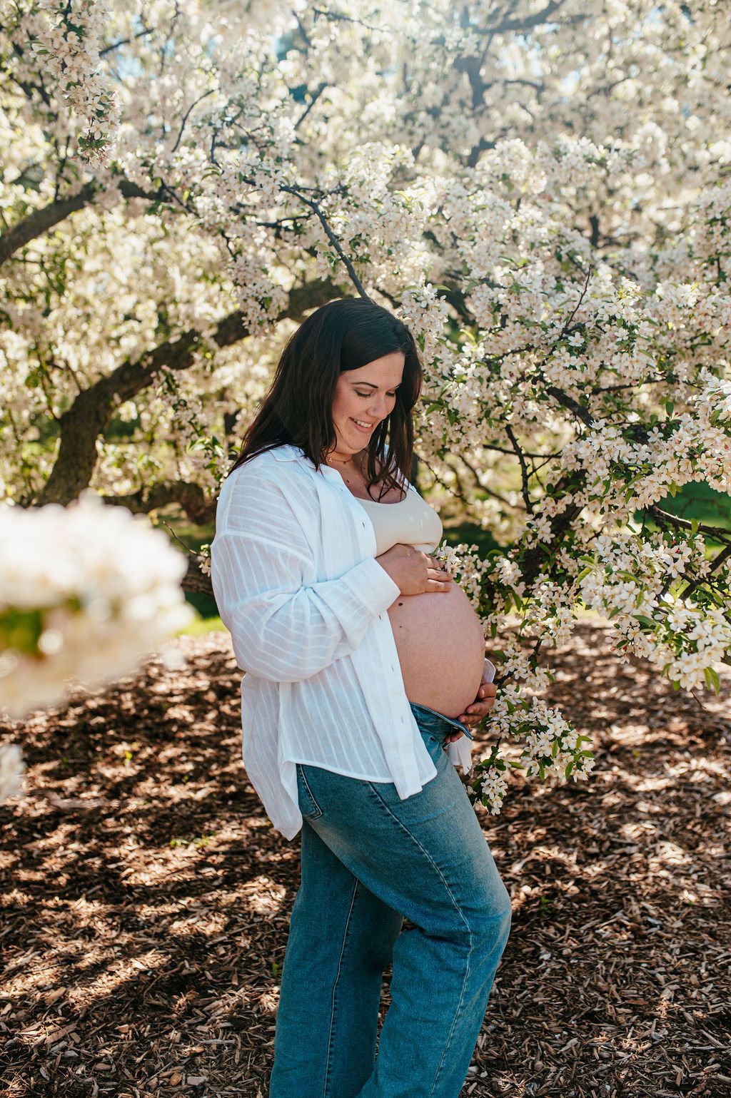 Pregnant person in a white shirt and jeans standing under blooming trees, looking at a phone.