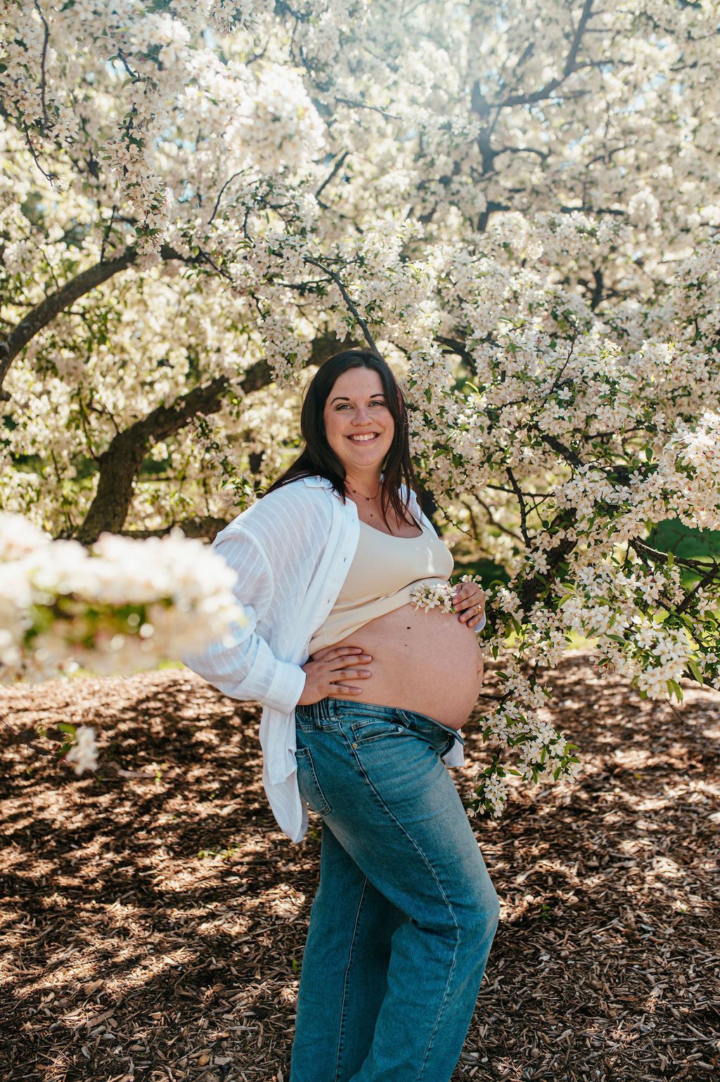 Pregnant woman smiling in a blooming orchard, wearing a white top and jeans.