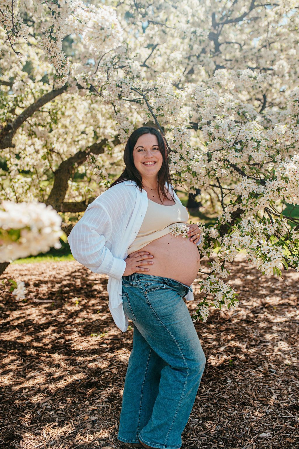 Pregnant person in a white cardigan and jeans standing under blooming trees outdoors