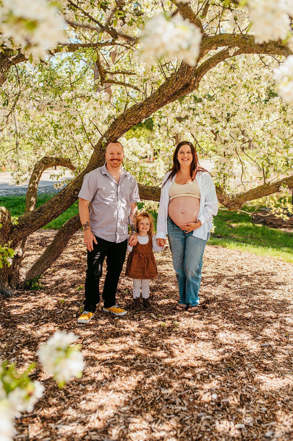 Three people standing under blooming tree branches in a sunlit park, smiling at the camera.