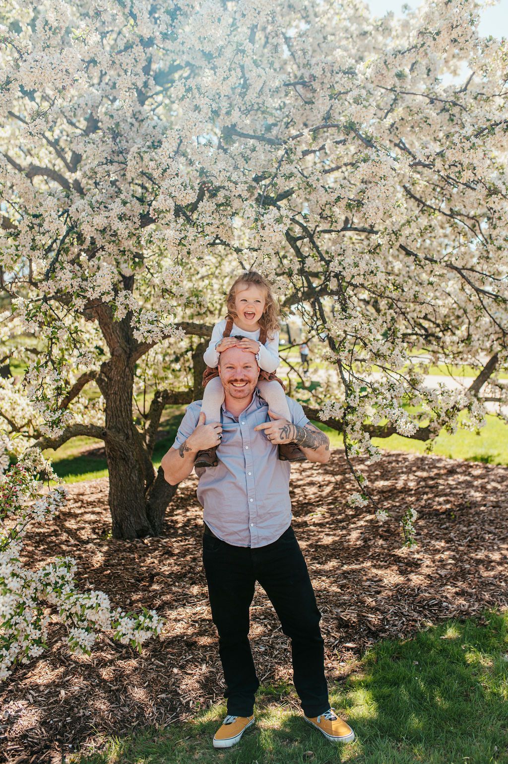 Person carrying a smiling child on shoulders under a blooming tree in a sunny garden