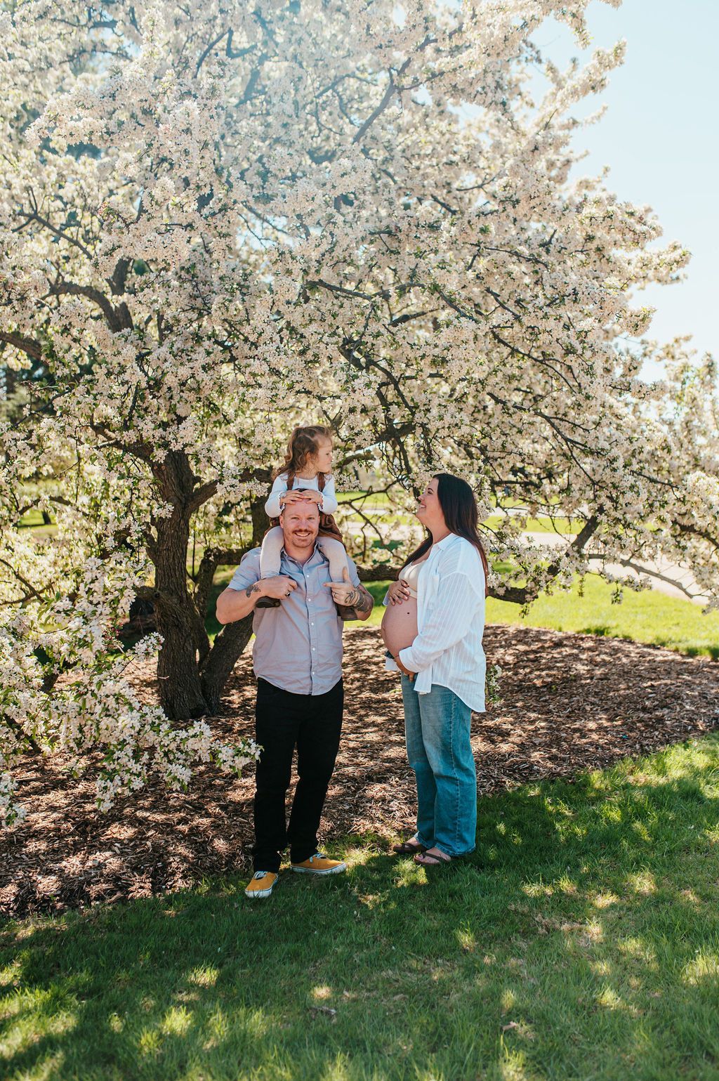 Three people standing under a blooming tree in a sunny park, one holding a child.