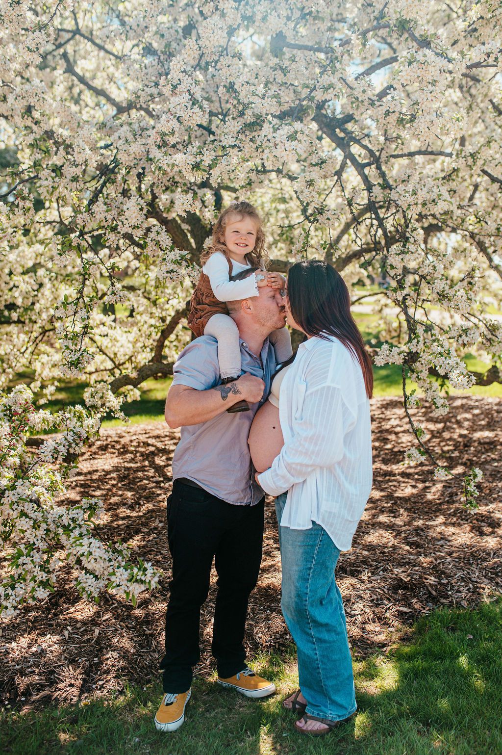 Three people standing under blooming trees, one holding a small child in a sunlit orchard.