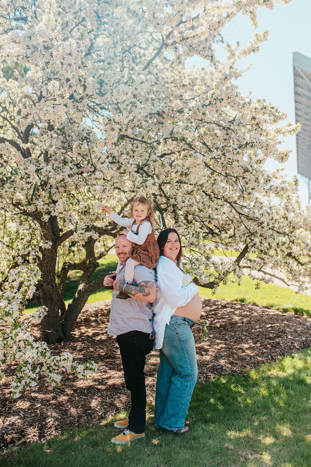 Three people posing under a blooming white tree in a sunny park