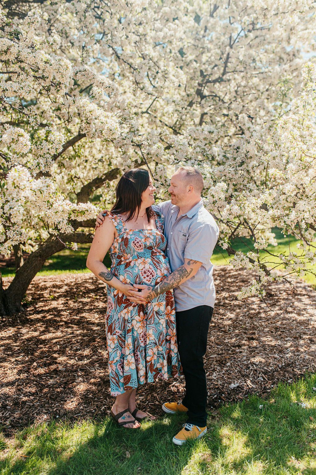 Couple standing together under blooming white tree blossoms in a sunlit park.