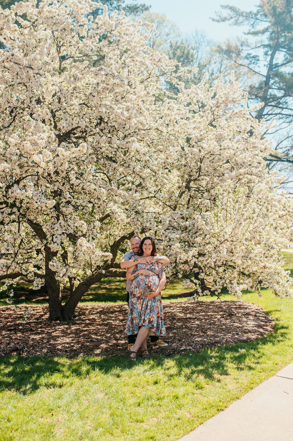 Person in a floral dress standing under a blooming tree in a sunny park