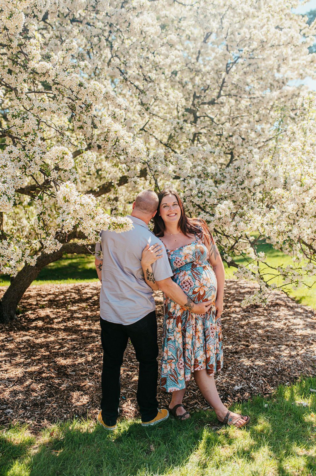 Couple embracing under a blooming white tree in a sunny garden, smiling at the camera.