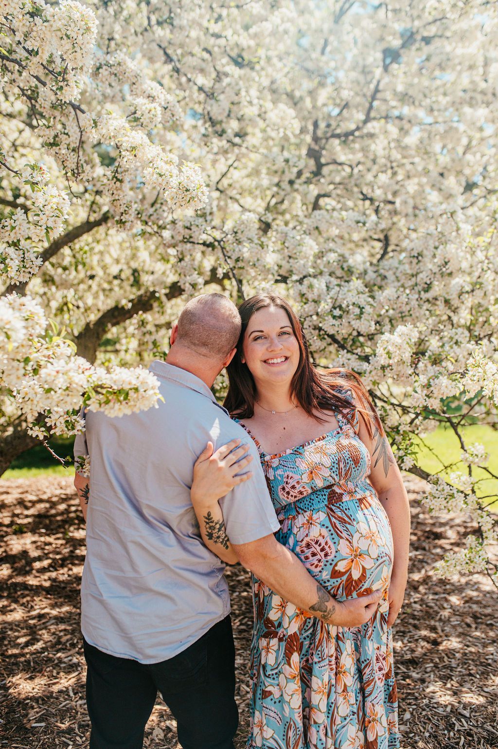 Couple standing together under blooming white blossoms, smiling in a sunlit garden.