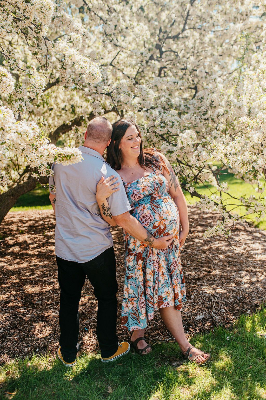 Pregnant couple standing under blooming tree, embracing and smiling outdoors