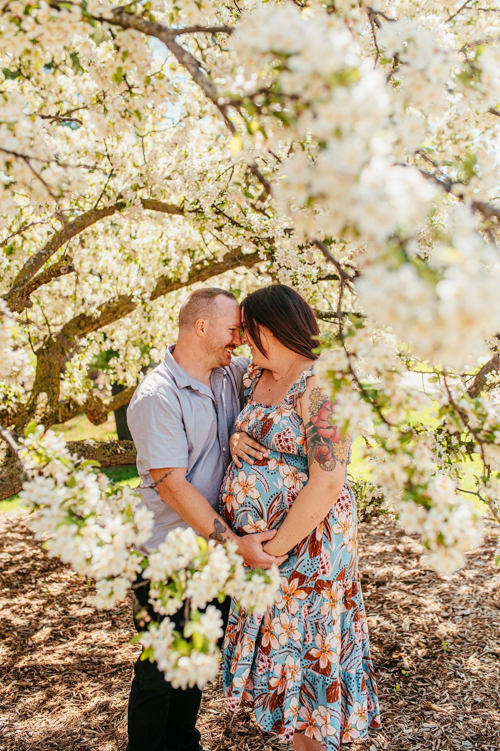 Couple standing close under blooming white blossoms, sharing a tender moment outdoors.