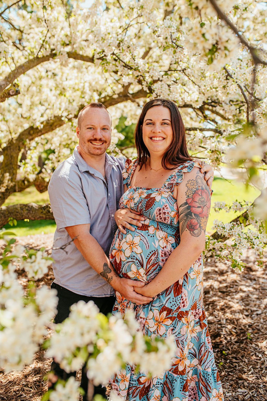 Couple embracing under blooming white blossoms in a sunlit garden, woman in floral dress holding a baby bump