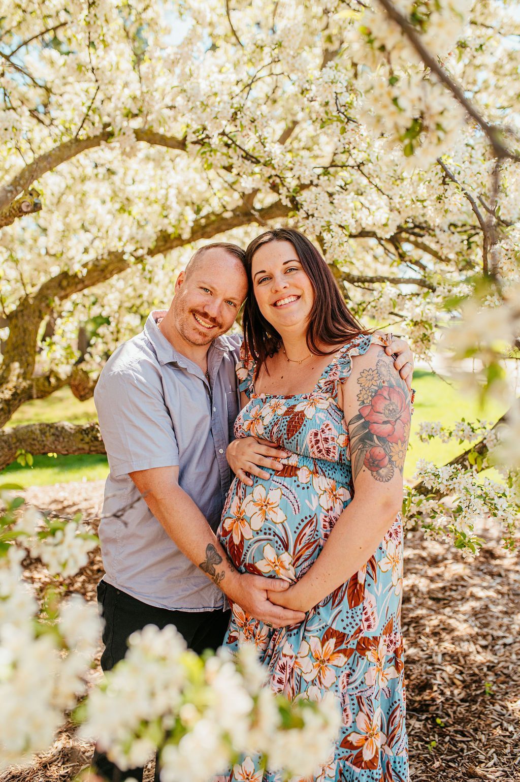 Smiling couple standing under blooming trees, holding a pregnant belly in a sunlit garden.