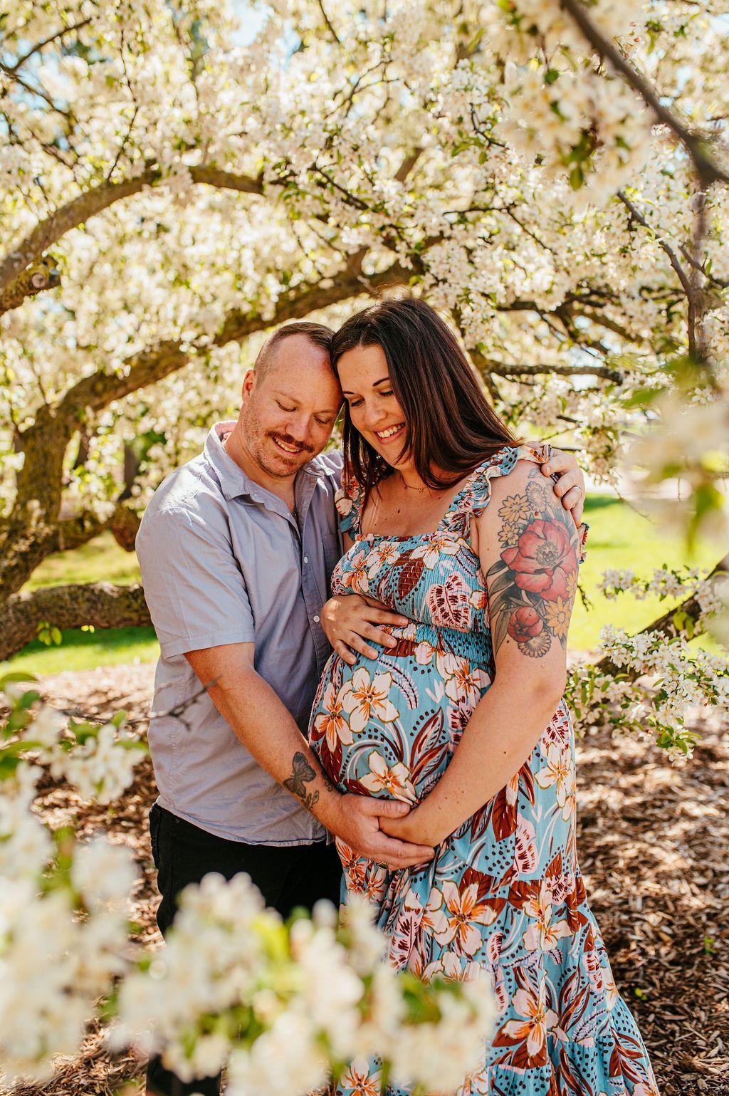 Couple embracing under blooming blossoms, with the woman cradling a baby bump in a floral dress.