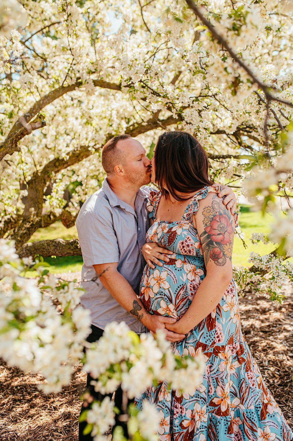 Couple embracing and kissing under blooming white blossoms, woman in floral dress and man in blue shirt