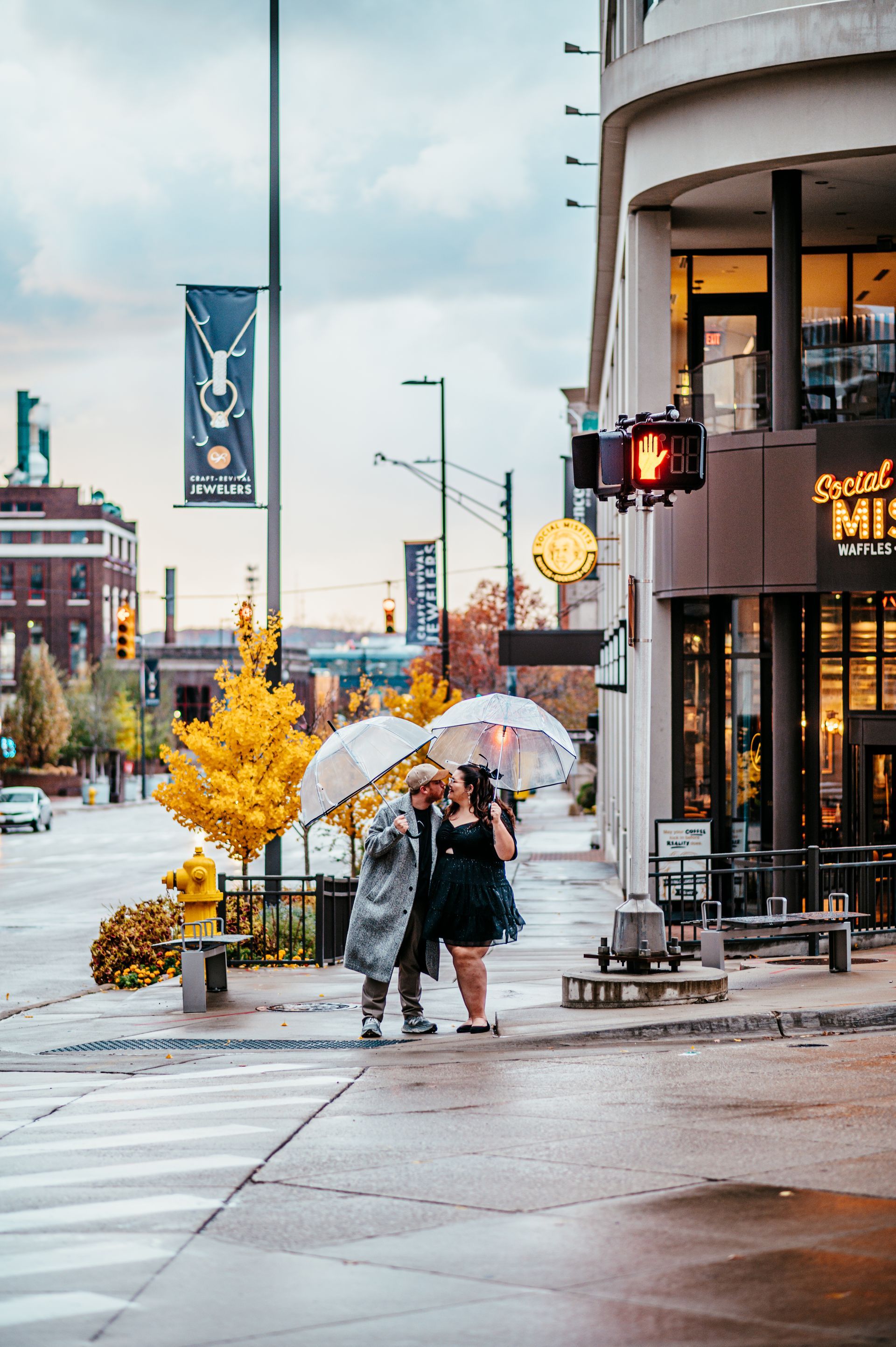 Rainy city sidewalk with two people under umbrellas near a lit storefront and yellow trees
