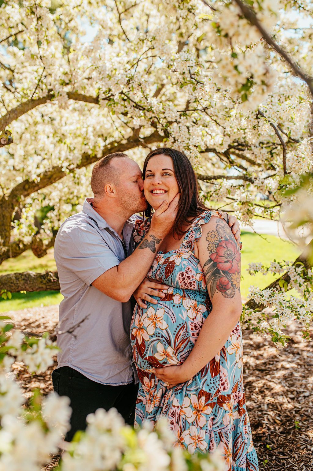 Couple standing under blooming white blossoms, smiling and embracing in a sunny garden.