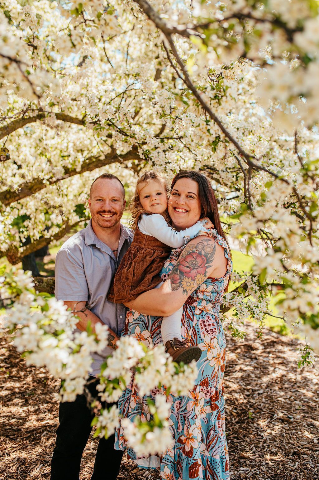 Family of three smiling under blooming white flowers outdoors, with parents holding a child.
