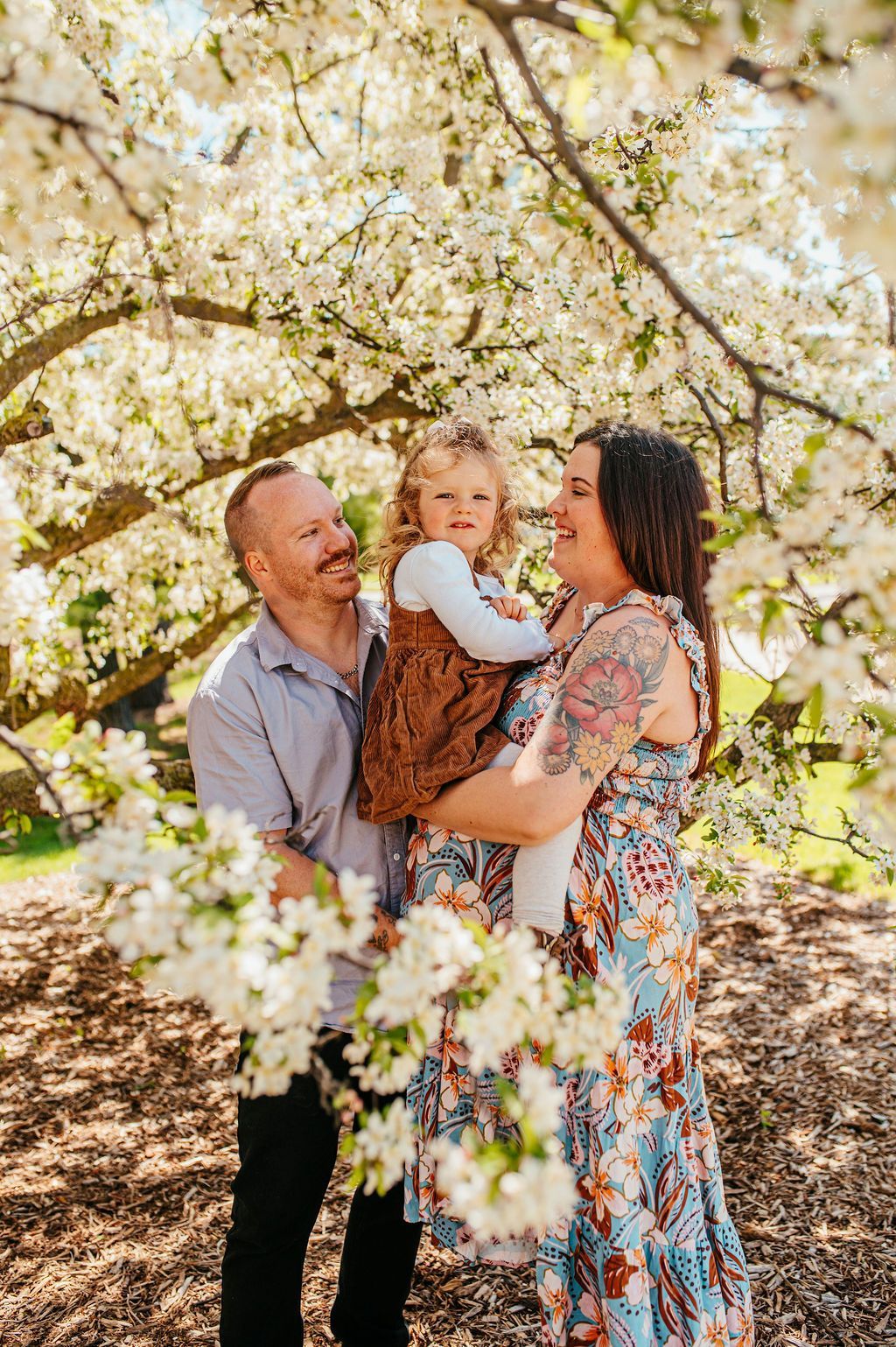 Family standing under blooming white blossoms, holding a toddler in a floral dress outdoors.