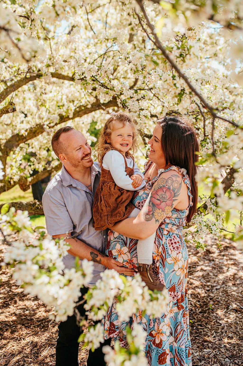 Family of three smiling in a blooming orchard, with parents holding a child under white blossoms.