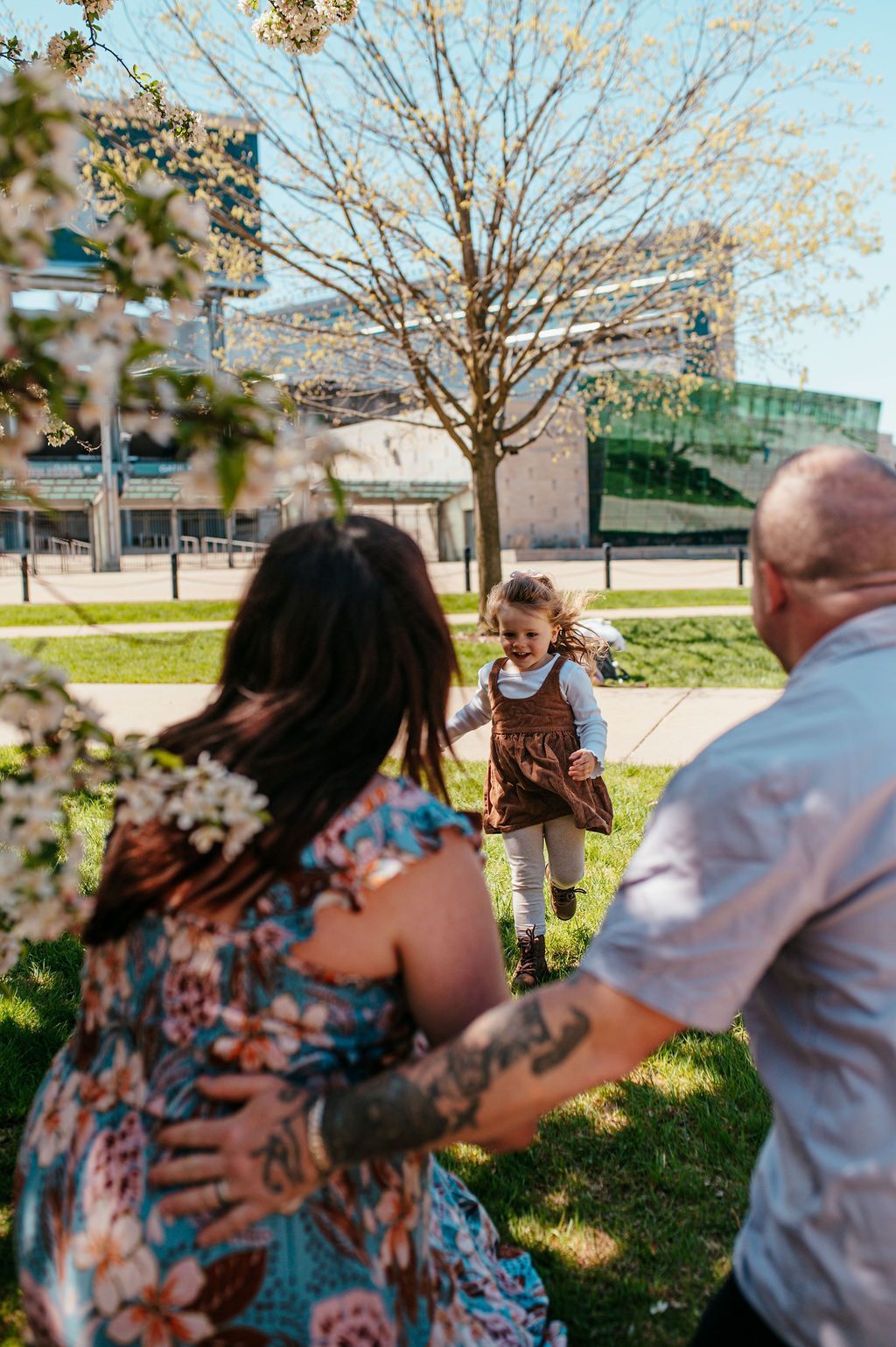 Two adults in a park reaching toward a child standing on the grass near a tree and buildings