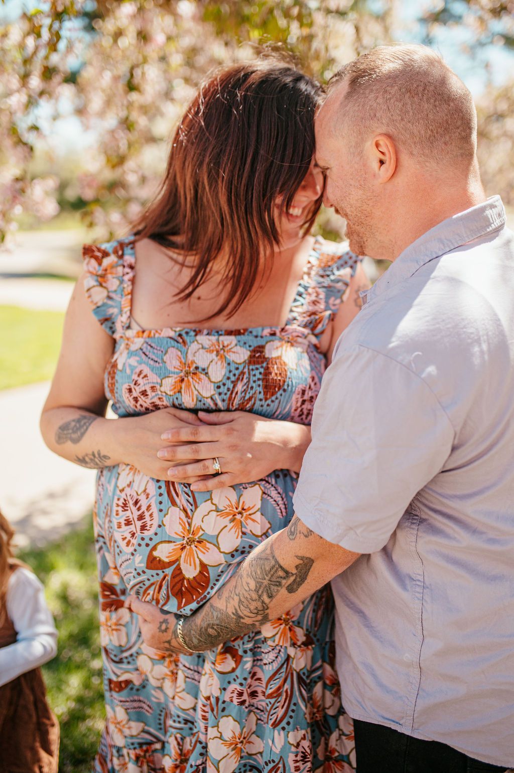 Couple embracing outdoors in a sunlit park, with the woman in a floral dress.