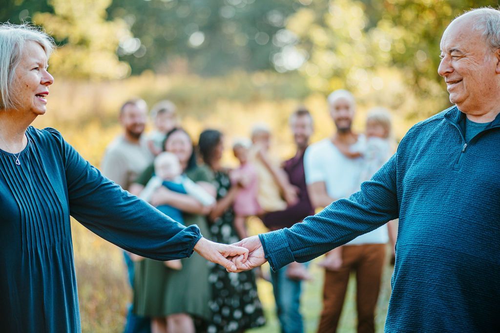Two people holding hands and smiling outdoors, with a group of people blurred in the background