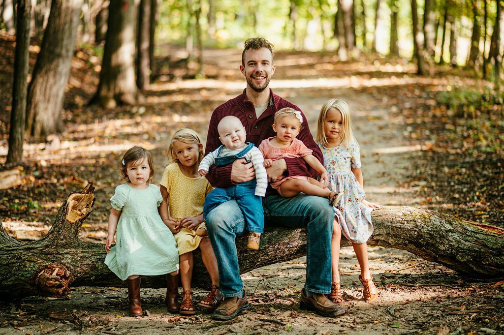 Family seated on a bench in a sunlit wooded park, with children gathered around a smiling man.