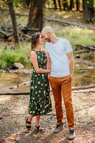Two people standing close together by a creek in a wooded area, smiling and touching foreheads.