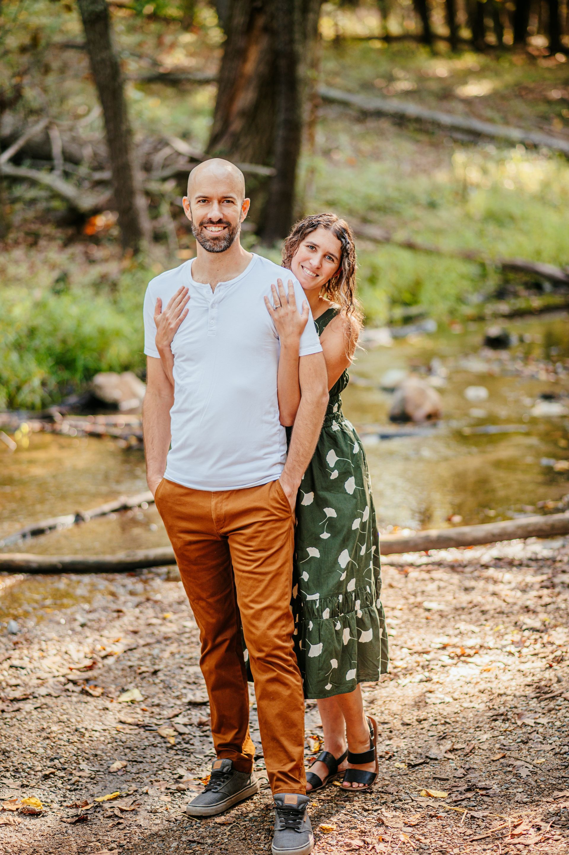 Couple standing by a creek in a wooded area, with the woman hugging the man from behind.