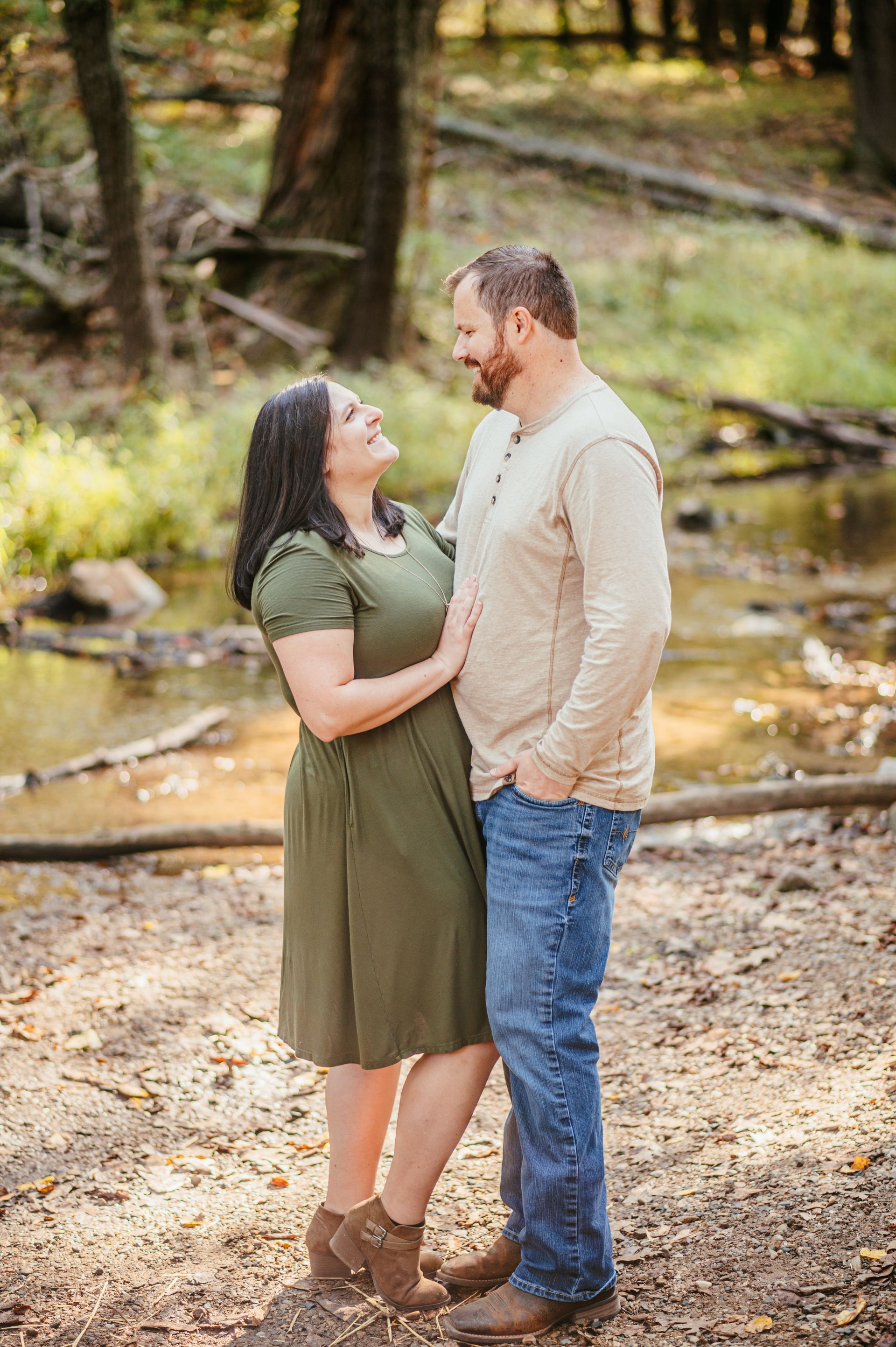 Couple standing face to face outdoors by a stream, smiling in a wooded setting.