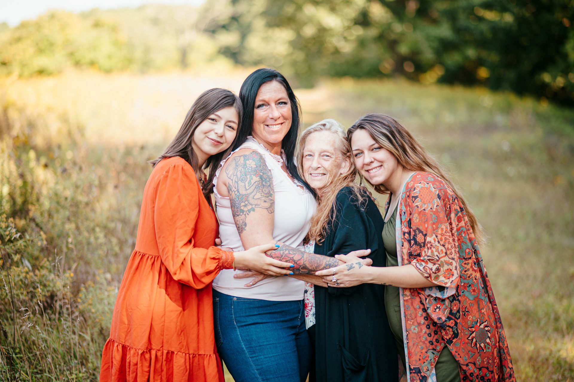 Four women in colorful dresses hugging and smiling in a sunlit field.