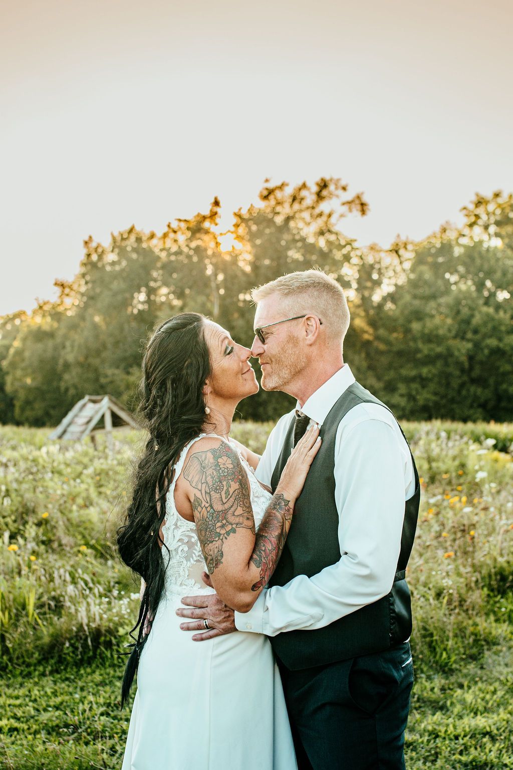 Couple embracing and kissing in a sunlit outdoor field at sunset, with trees in the background.