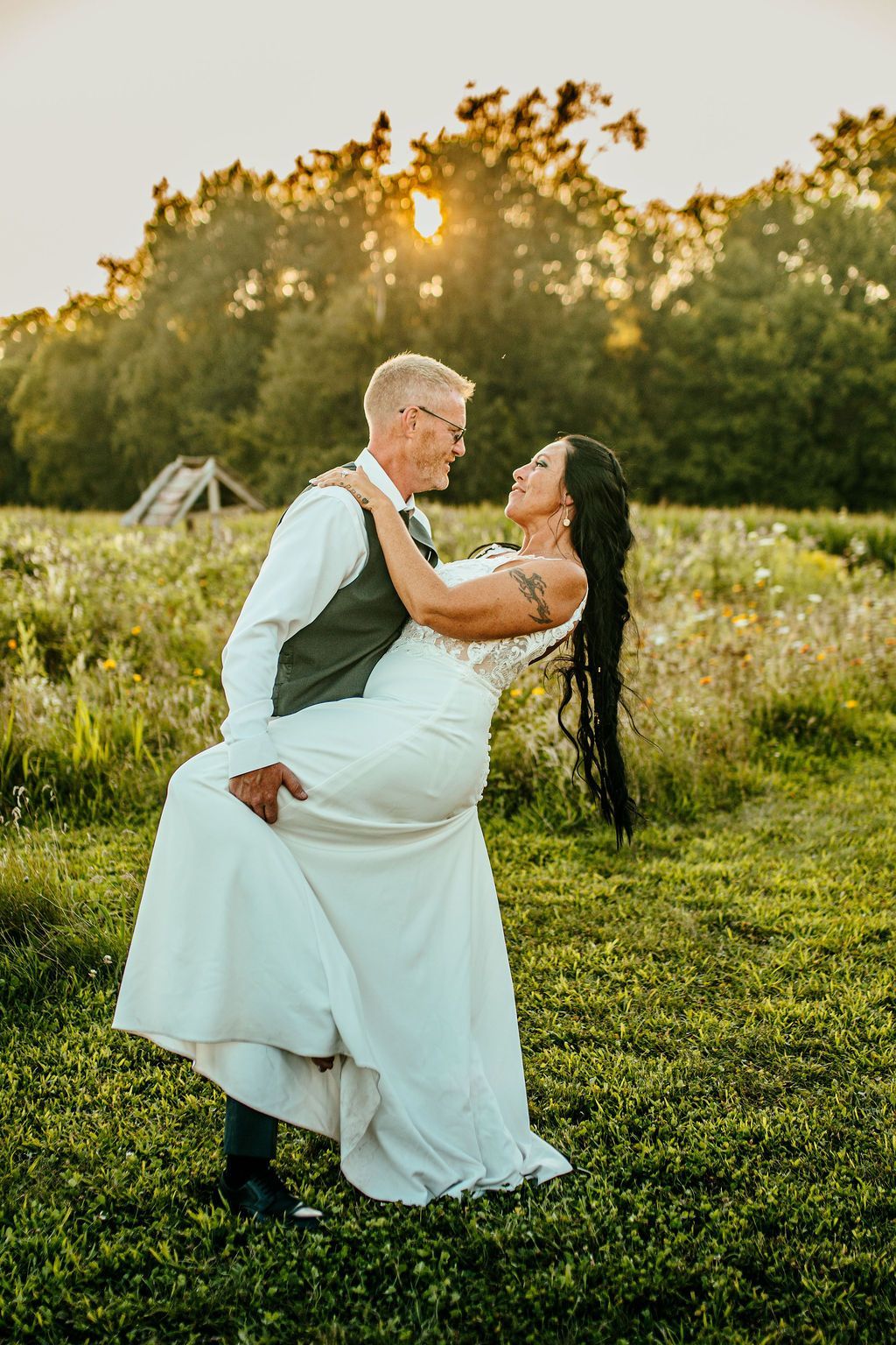 Couple in white wedding attire embracing in a sunlit grassy field at sunset.