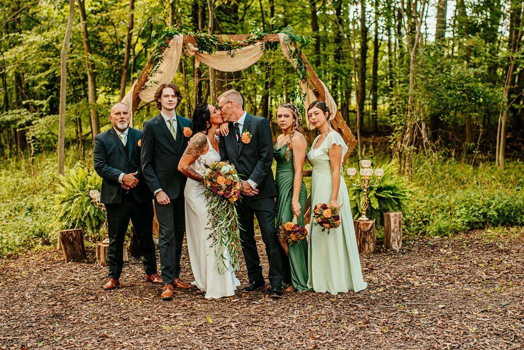 Wedding party posing outdoors in a wooded clearing, with bouquets and formal attire.