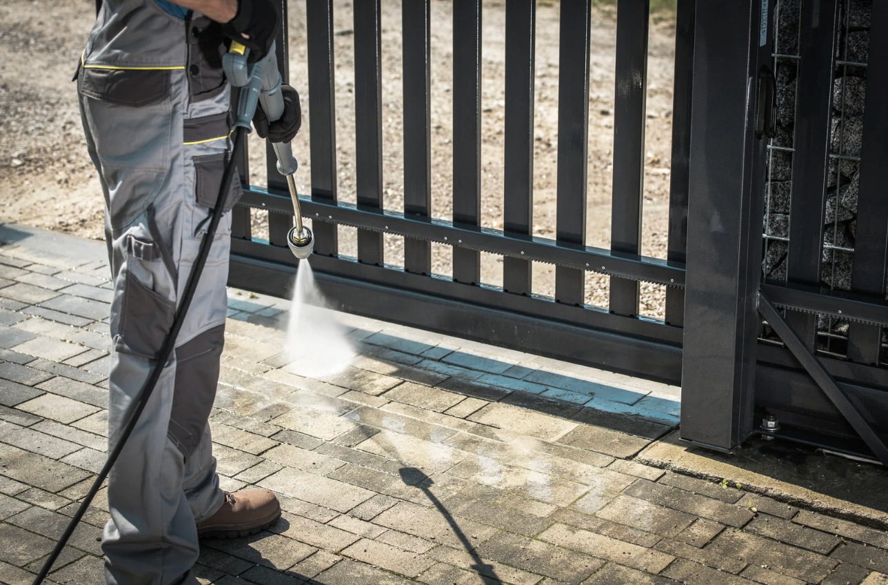 Worker pressure-washing pavement beside a black metal gate with a white spray plume
