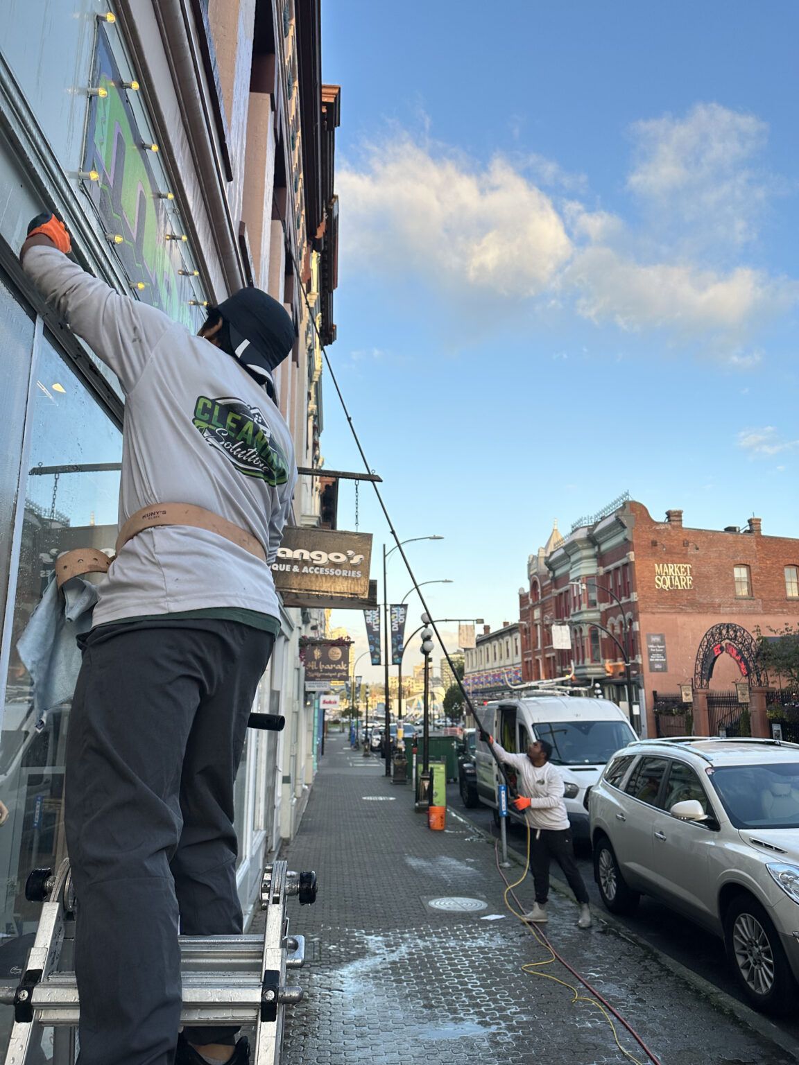 Worker on a ladder fixing a building facade on a wet city street with parked cars and blue sky.
