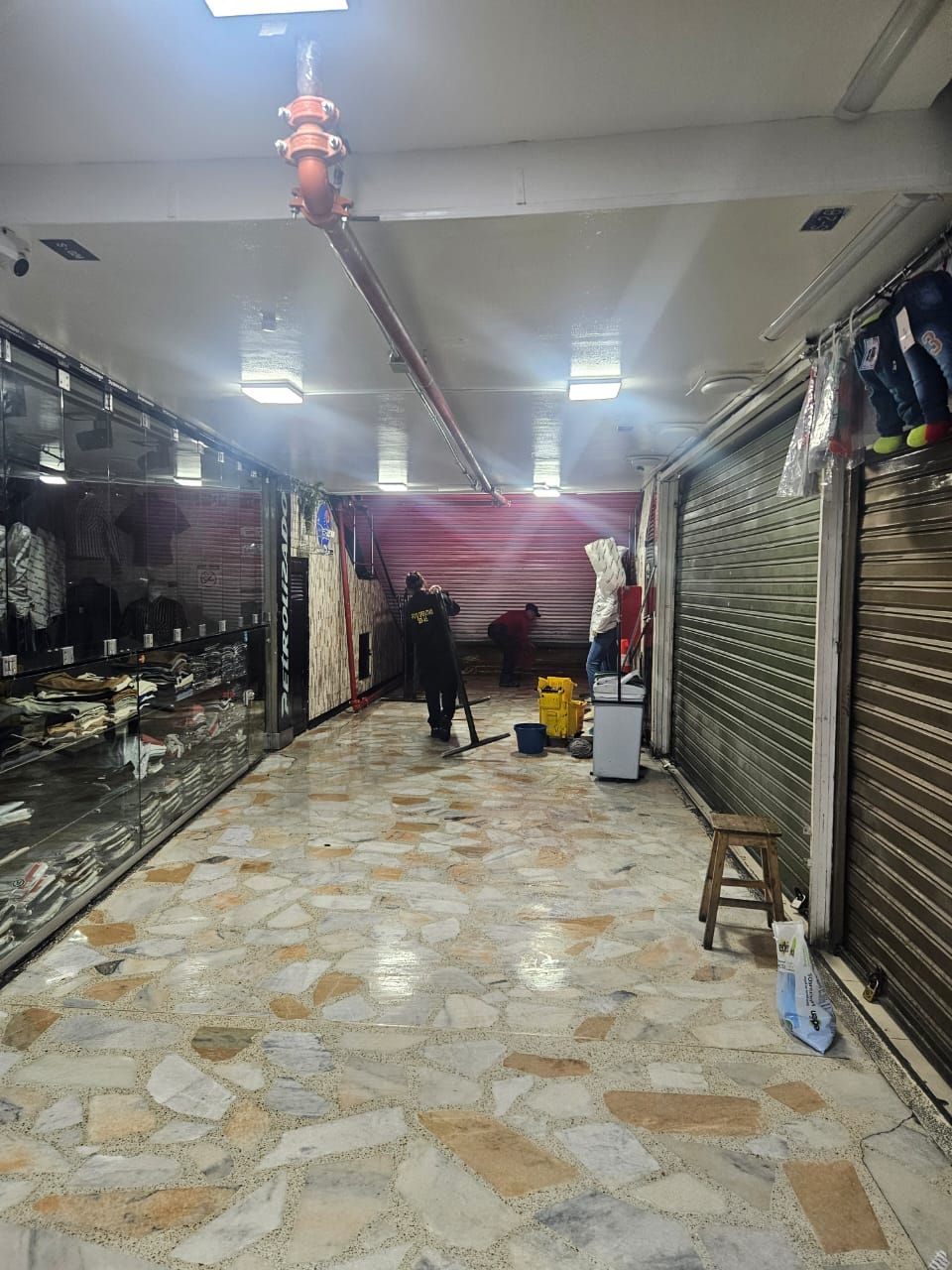 Pasillo de mercado interior con persianas metálicas cerradas, una persona limpiando y suelo de baldosas estampadas.